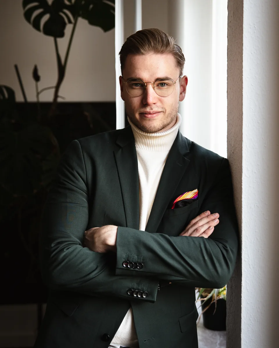 Nicholas Bode mit verschränkten Armen am Fenster, Blick in die Kamera, dunkelgrünes Sakko und heller Rollkragenpullover