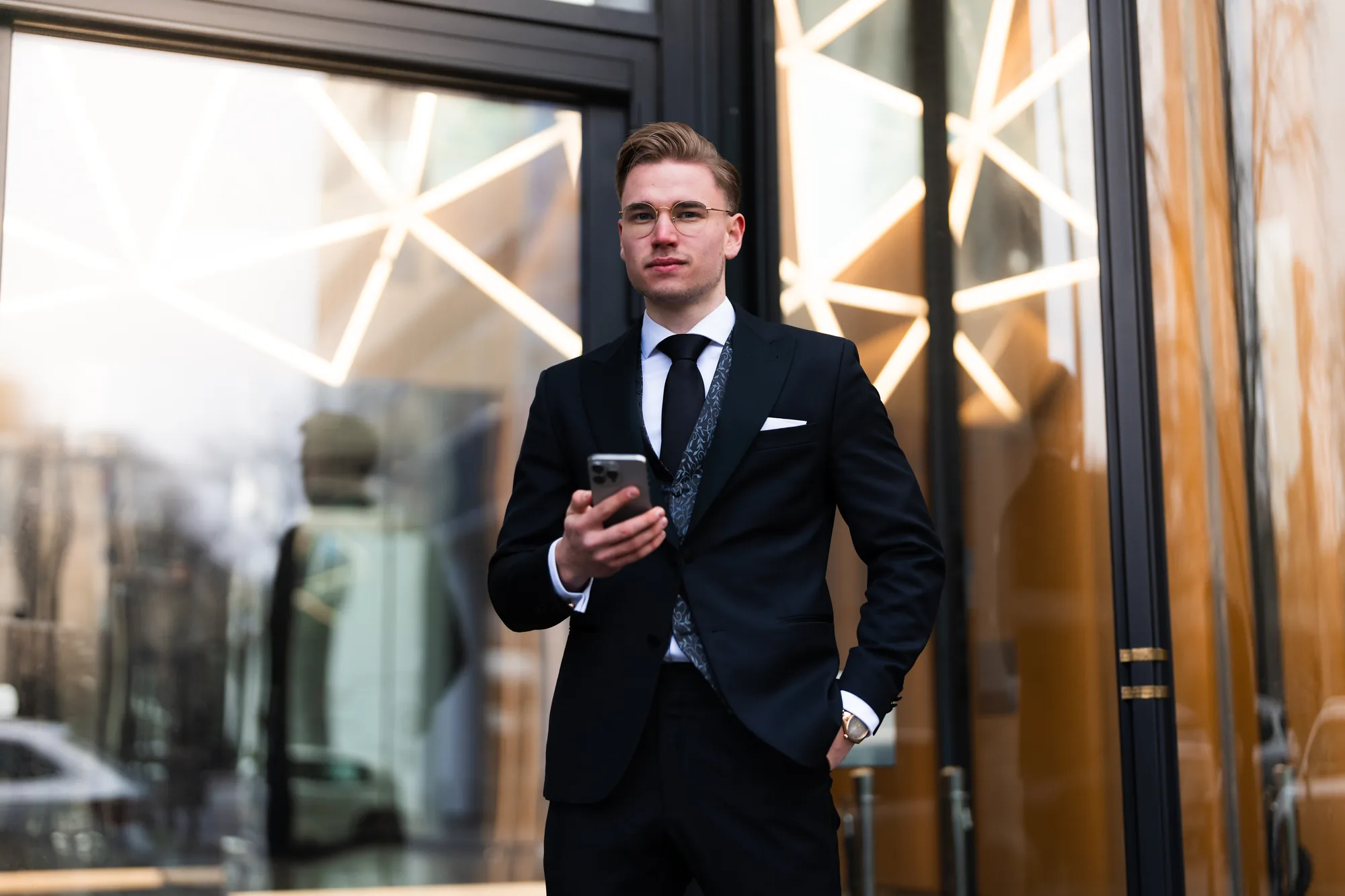 Nicholas Bode im dunklen Anzug mit Brille vor einer Glasfassade in Frankfurt, das Smartphone in der Hand